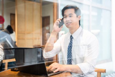 Beyaz gömlekli ve mavi kravatlı bir adam laptopuyla meşgul ve cep telefonuyla konuşuyor.