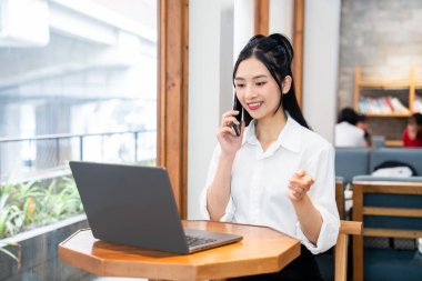 Bir kadın telefonda konuşurken gülümsüyor. Parlak bir çalışma alanında dizüstü bilgisayarla.