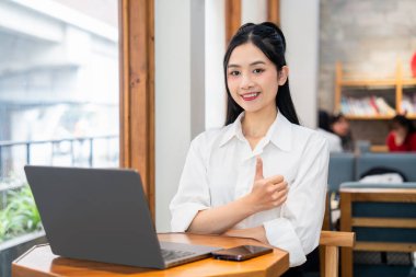 Bir kafede dizüstü bilgisayarında çalışan gülümseyen genç bir kadın onaylı bir jest yapıyor.