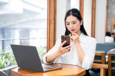 Genç bir kadın kafede telefonunu kullanıyor. Önünde dizüstü bilgisayarı var. Düşünceli biri.