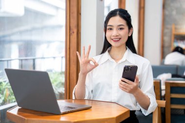 Asyalı bir kadın bir kafede dizüstü bilgisayar kullanarak oturuyor ve elinde bir akıllı telefon tutuyor.