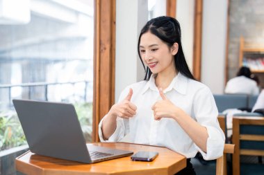 Neşeli genç bir profesyonel, bir dizüstü bilgisayarla çalışıyor ve ışıklı bir kafe ortamında onay veriyor.