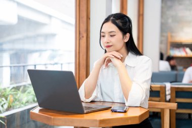 Genç bir kadın bilgisayarlı ve telefonlu masada oturuyor. Uzaktan kumandalı parlak bir kafede düşünceli görünüyor.