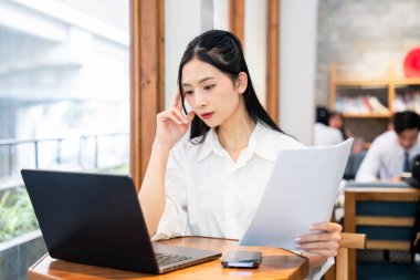 Profesyonel olarak giyinmiş genç bir kadın dizüstü bilgisayarı üzerinde çalışıyor ve elinde derin düşüncelere dalmış belgeler tutuyor.