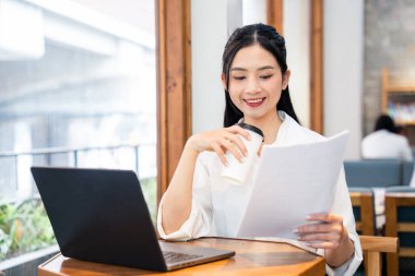 Gülümseyen Asyalı kadın belgeleri inceliyor ve kahve fincanını elinde dizüstü bilgisayarla bir kafede tutuyor.