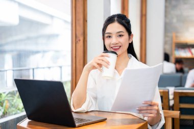 Gülümseyen bir kadın evrak işleriyle uğraşırken kahvesini yudumlar ve bir kafede dizüstü bilgisayarıyla çalışır.