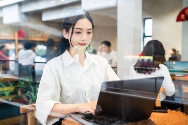 Kadın, kafede dizüstü bilgisayarda çalışırken masada oturur ve diğer müşteriler vitrinden görülebilir.