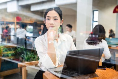 Kafede çalışan düşünceli bir kadın masadaki dizüstü bilgisayardan baktığında şehir manzarasını yansıtıyor.