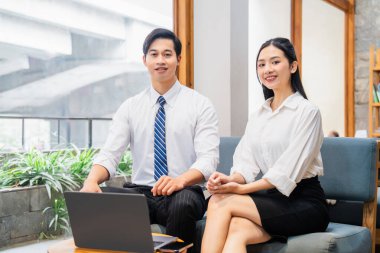 Gülümseyen adam ve kadın profesyonel olarak giyinip dizüstü bilgisayar kullanıyor ve mavi bir koltukta oturuyorlar.