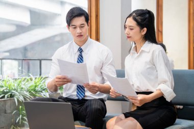 İki iş arkadaşı, modern bir ofis ortamında dizüstü bilgisayarla fikir alışverişinde bulunan belgeleri analiz ediyor.