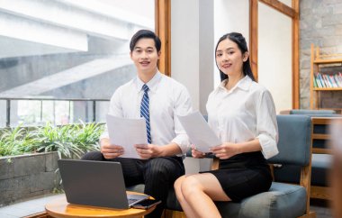 İki Asyalı meslektaşı ellerinde belgelerle gülümsüyor. Masada dizüstü bilgisayarlarıyla oturuyorlar.