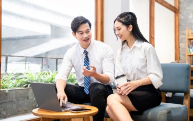 Asyalı bir erkek ve kadın profesyonel olarak dizüstü bilgisayarlarını güneşli bir kafe veya ofis alanında gözden geçiriyorlar.