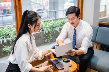 İki meslektaş, Asyalı bir kadın ve bir adam laptopları ve gülümseyen belgelerle bir kafede birlikte çalışıyorlar.