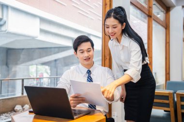 Havai bir ofiste birlikte çalışan iki genç profesyonel pencere kenarındaki evrakları gözden geçiriyorlar.