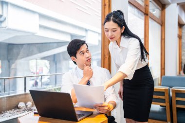 İki iş adamı parlak pencereli bir alanda danışmanlık yapıyor. Adam belgeleri incelerken dalgın görünüyor.