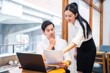 İki meslektaş modern bir düzende işbirliği yapıyorlar. Bir erkek ve kadın, pencere ve dizüstü bilgisayarın yanında kağıtları gözden geçiriyorlar.