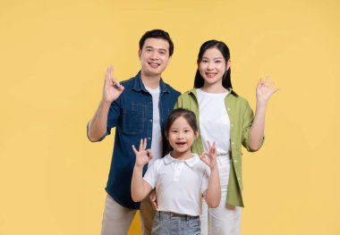 Üç kişilik mutlu bir aile, parlak sarı bir fon önünde poz veriyor ve gülümsüyor.