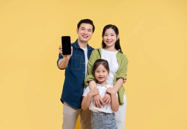Gülümseyen bir aile sarı arkaplan önünde poz veriyor. Baba reklam için cep telefonu tutuyor.