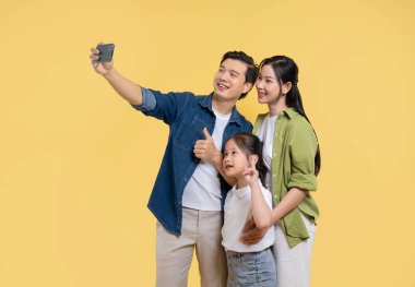 Gülümseyen baba, anne ve kız selfie için poz veriyor. Sade sarı bir arkaplana karşı. Mutlu görünüyorlar.