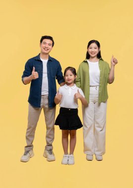 Üç kişilik mutlu bir aile, sıradan giyinmiş ve başparmak kaldıran sarı bir zemine karşı dururlar.