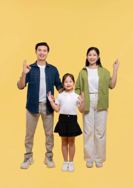 Üç kişilik aile, sarı bir zemine karşı durup gülümserken el hareketlerini bir arada tutuyorlar.