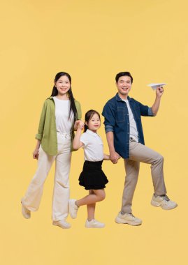 Üç kişilik mutlu bir Asyalı aile elinde kağıttan bir uçak tutan sarı bir arka planda duruyor.