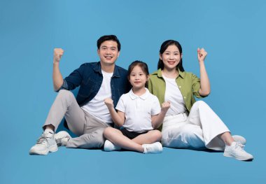 Üç kişilik gülümseyen Asyalı bir aile bir baba ve bir kız çocuğu mavi bir zemine karşı sevinçle otururlar.