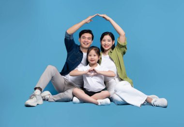 Üç kişilik mutlu bir aile, mavi bir zeminde el ele tutuşmuş bir çatı ve kalp şeklinde poz veriyor.