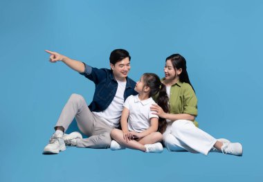 Gülümseyen üç kişilik bir aile mavi bir arka planda oturup dışa bakıyor ve işaret ediyor.