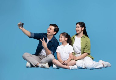 Üç kişilik neşeli bir aile, canlı mavi bir arka planda mutluluk saçan bir selfie için poz veriyor.