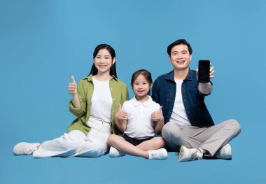 Gülümseyen aile pozları başparmakları havada baba mavi arka planda bir akıllı telefon tutuyor.