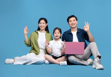 Üç kişilik bir aile gülümsüyor ve mavi bir arkaplanda oturuyor. Bilgisayarı paylaşıyor ve işaret ediyor. Tamam.