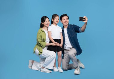 Üç kişilik mutlu bir aile akıllı telefonuyla çekilen bir fotoğraf için mavi bir arka plan karşısında gülümsüyor.