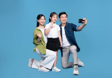 Üç kişilik gülümseyen bir aile, mutluluk ve birlik saçan açık mavi bir arkaplana karşı selfie için poz verir.