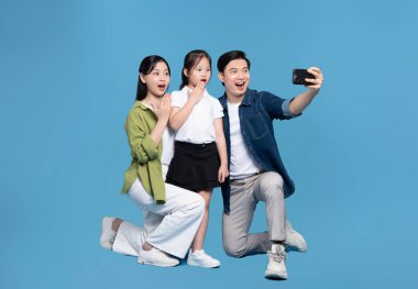 Şaşırmış üç kişilik bir aile, stüdyo ortamında mavi arka planda telefonla fotoğraf çekiyor.