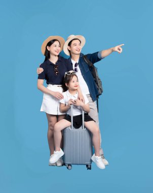 Üç kişilik bir aile seyahat kıyafetleri içinde parlak mavi bir arkaplana karşı bavulla poz veriyor.