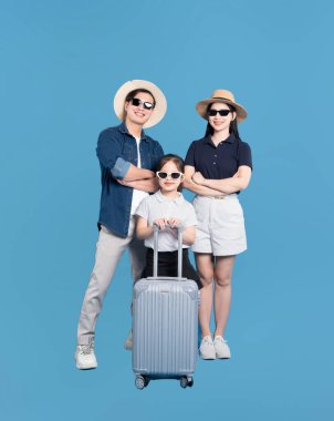 Üç kişilik aile güneş gözlüğü takıyor ve mavi arka planda bavulla poz veriyor. Gülümsüyor.