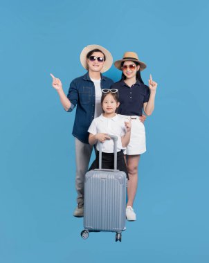 Üç kişilik gülümseyen bir aile, parlak mavi bir arkaplanı işaret eden bavullarıyla tatile çıkacak.