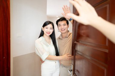 Gülümseyen bir çift, evlerinin kapısında durur ve görünmeyen bir insanı selamlar.