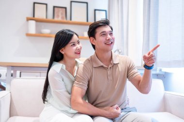 Çift beyaz koltukta oturuyor ve adam kameranın dışında bir şeye işaret ederken gülümsüyor.