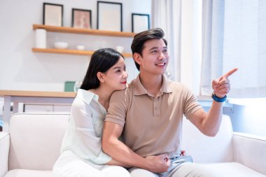 Bir çift kanepeye oturmuş ileriye bakıyor ve adam parmağını gösterip gülümsüyor.