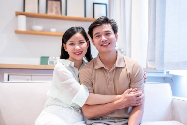 Mutlu bir Asyalı çift, aydınlık, modern bir oturma odasındaki koltukta otururken sıcacık bir şekilde gülümsüyor.