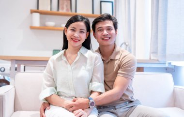 Mutlu bir çift, parlak, modern bir oturma odasındaki koltukta oturup kameraya gülümsüyor.