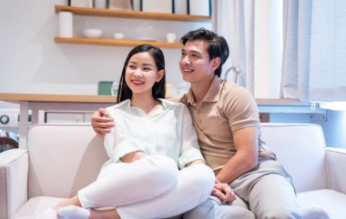 Asyalı bir çift, parlak, modern bir oturma odasına bakarken koltukta gülümsüyor.