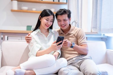 Gülümseyen bir çift, parlak minimalist bir oturma odasındaki akıllı telefona bakarak kanepede rahatlar.