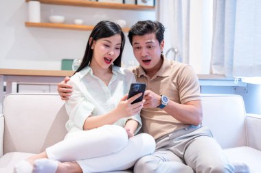 Asyalı bir çift, modern bir evde beyaz bir kanepede birlikte telefon izlerken şok olmuş görünüyor.