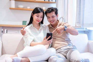 Mutlu bir çift evdeki kanepede rahatlar. Mobil bir cihazın içeriğine bakar ve anı paylaşır.