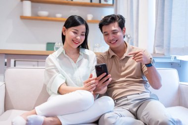 Kanepede yan yana oturan genç bir çift cep telefonunu izliyor ve gülümsüyor.