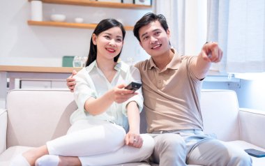 Oturma odasında birlikte televizyon izlerken krem renkli bir koltukta dinlenen mutlu bir çift.