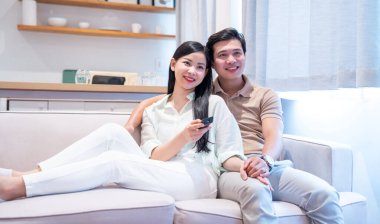 Oturma odasındaki kanepede uzanan mutlu bir çift evde birlikte televizyon izliyor ve gülümsüyor.